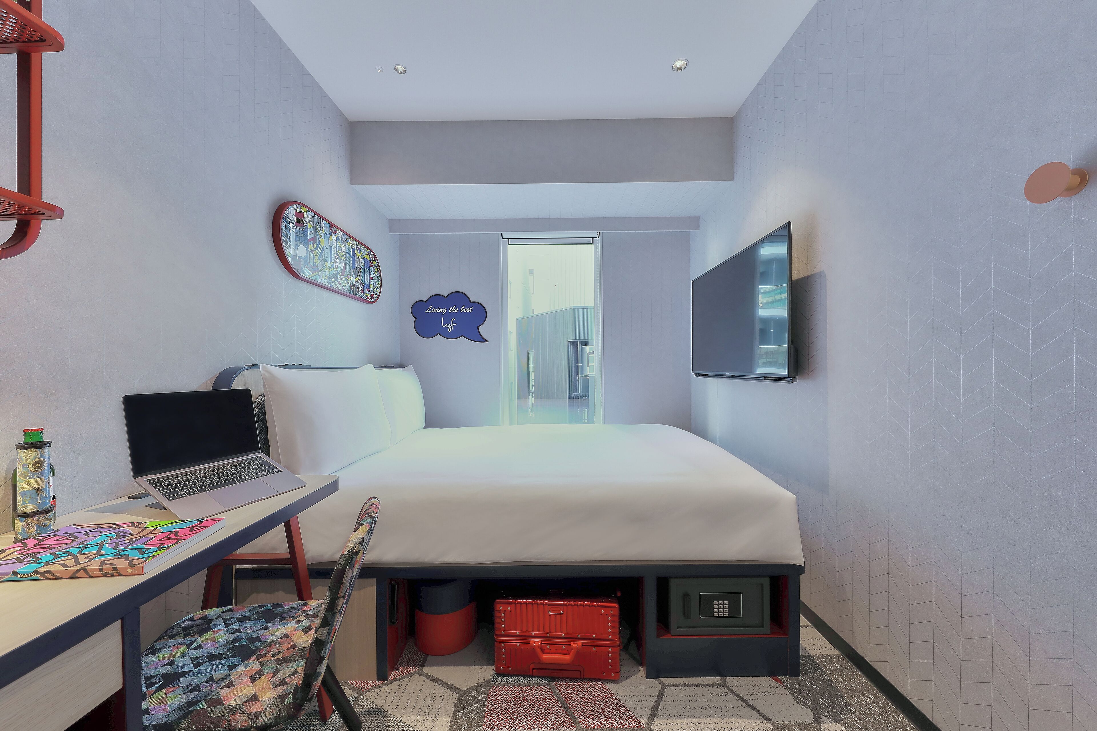 Habitación de hotel pequeña con una cama, un escritorio con una computadora portátil, una silla, un televisor montado en la pared y una ventana con vistas a la ciudad.