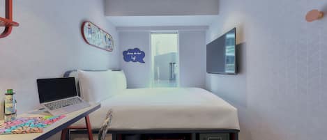 Habitación de hotel pequeña con una cama, un escritorio con una computadora portátil, una silla, un televisor montado en la pared y una ventana con vistas a la ciudad.