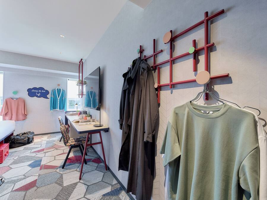 Habitación de hotel con cama, escritorio, silla, perchero con ropa y pared con diseño geométrico.