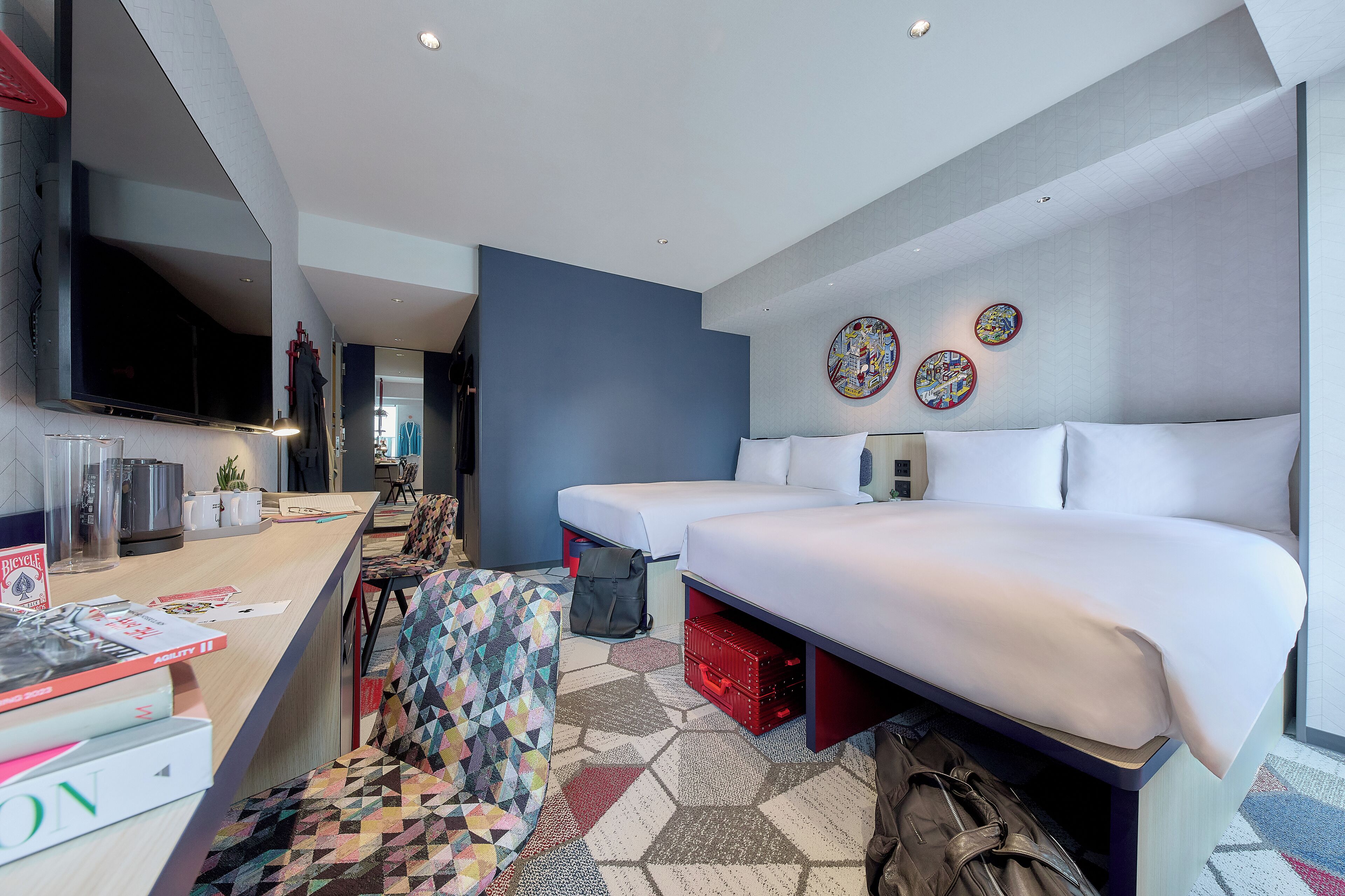 A modern hotel room with a large bed, a desk with a flat-screen TV, and a seating area with a colorful geometric-patterned chair.