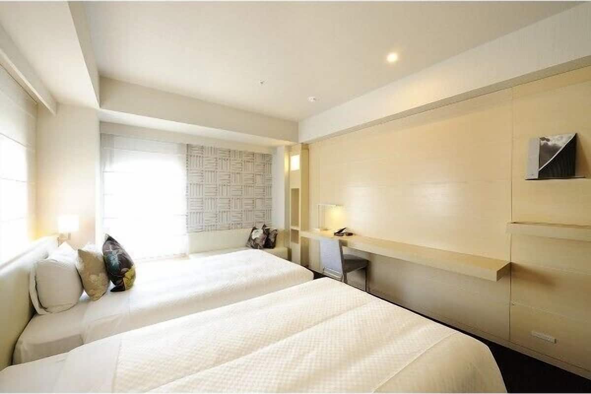 A hotel room with two beds, a desk, a chair, a lamp, and a wall-mounted TV.