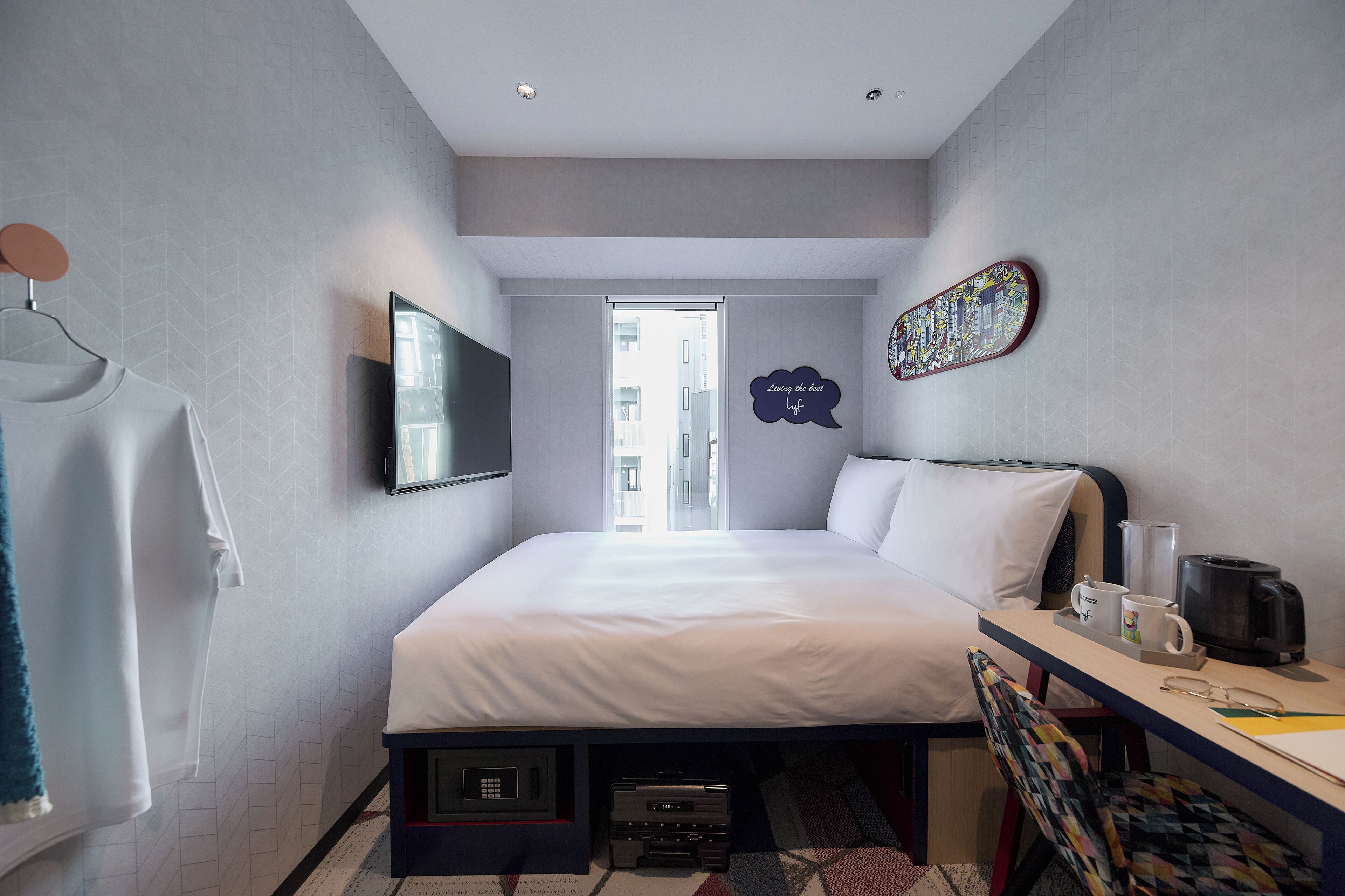 A modern hotel room with a bed, desk, and a view of the city through the window.