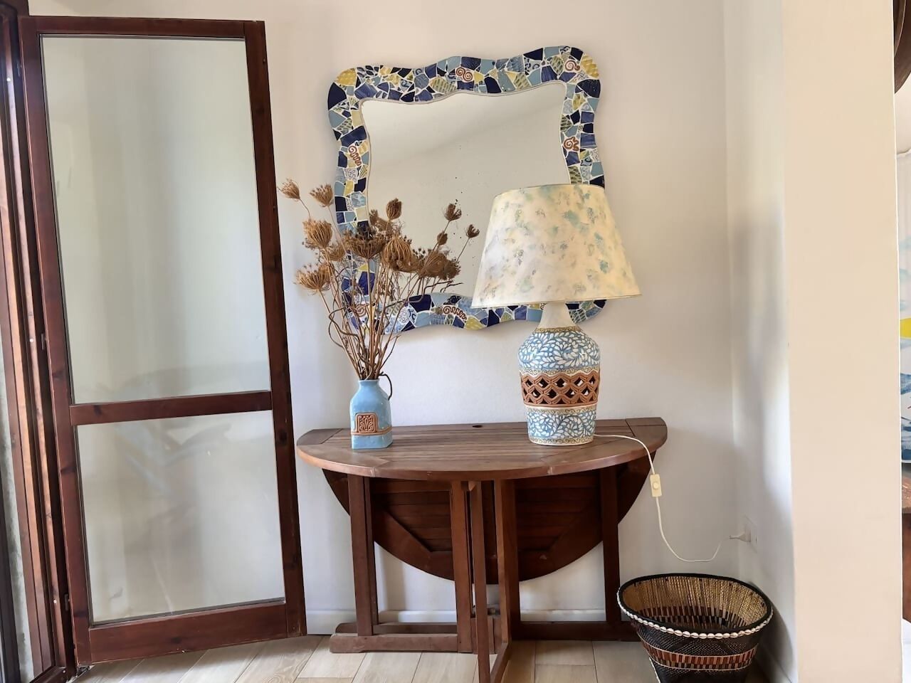 A room with a wooden table, a lamp, a vase with dried flowers, and a decorative mirror with a mosaic frame.