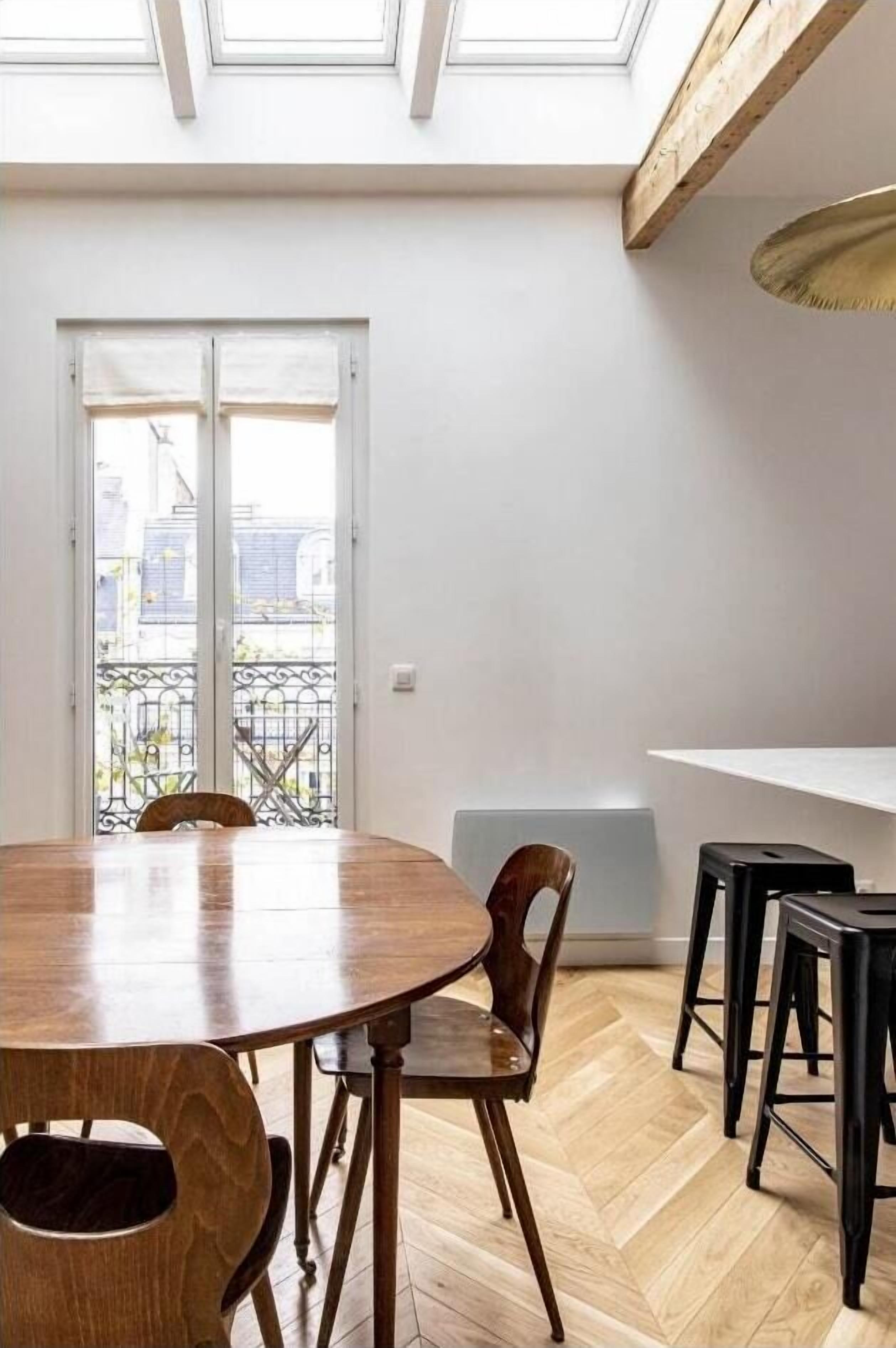 Een eethoek met een houten tafel en stoelen, een witte muur, en uitzicht door een glazen deur op een buitenbalkon.