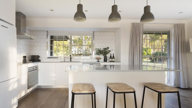 Une cuisine moderne avec des meubles blancs, un grand îlot central doté de tabourets, un éclairage suspendu et de grandes fenêtres avec des rideaux.