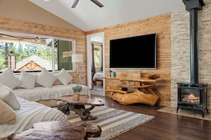 A cozy living room with a sofa, a flat-screen TV, a wood-burning stove, and a rustic wooden wall.