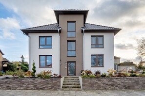 Ein zweigeschossiges Wohnhaus mit einem zentralen Eingang, einer Backsteinkonstruktion als Stützmauer und einem angelegten Garten.