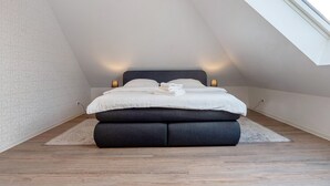 Une chambre avec un plafond en pente, un lit doté d’une tête de lit sombre, des lampes de chevet et une vue sur le ciel grâce à une lucarne.