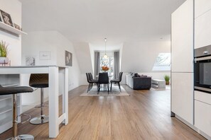 Eine moderne Küche mit weißen Schränken, einem Essbereich mit Tisch und Stühlen sowie einem Blick ins Freie durch ein Fenster.