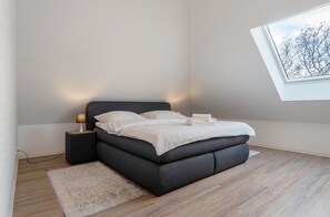Ein modernes Schlafzimmer mit einem dunkelbraunen Bett, einer Nachttischlampe, einem Fenster mit Blick auf Bäume und einem Holzboden.