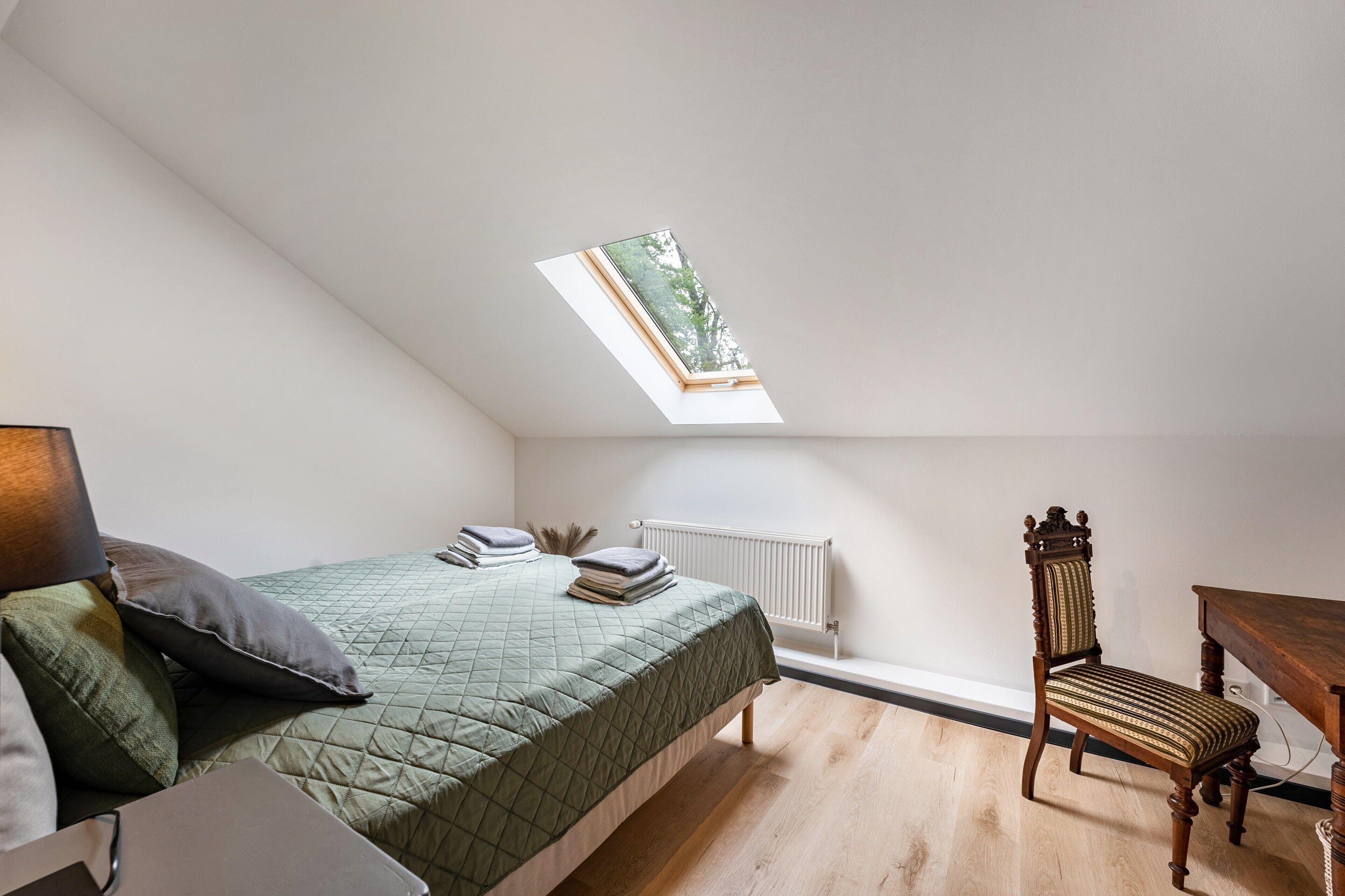 Une chambre avec un puits de lumière, un lit recouvert d’une courtepointe verte matelassée, une chaise en bois et une petite table.
