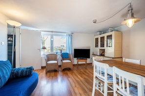 Quarto com uma mesa de jantar de madeira, duas cadeiras de vime, um sofá azul, uma televisão e um armário com portas de vidro.