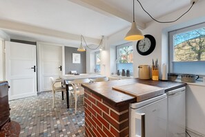 Een keuken met een tegelwand, witte kasten, een houten aanrechtblad, hanglampen, een klok en een raam met jaloezieën.