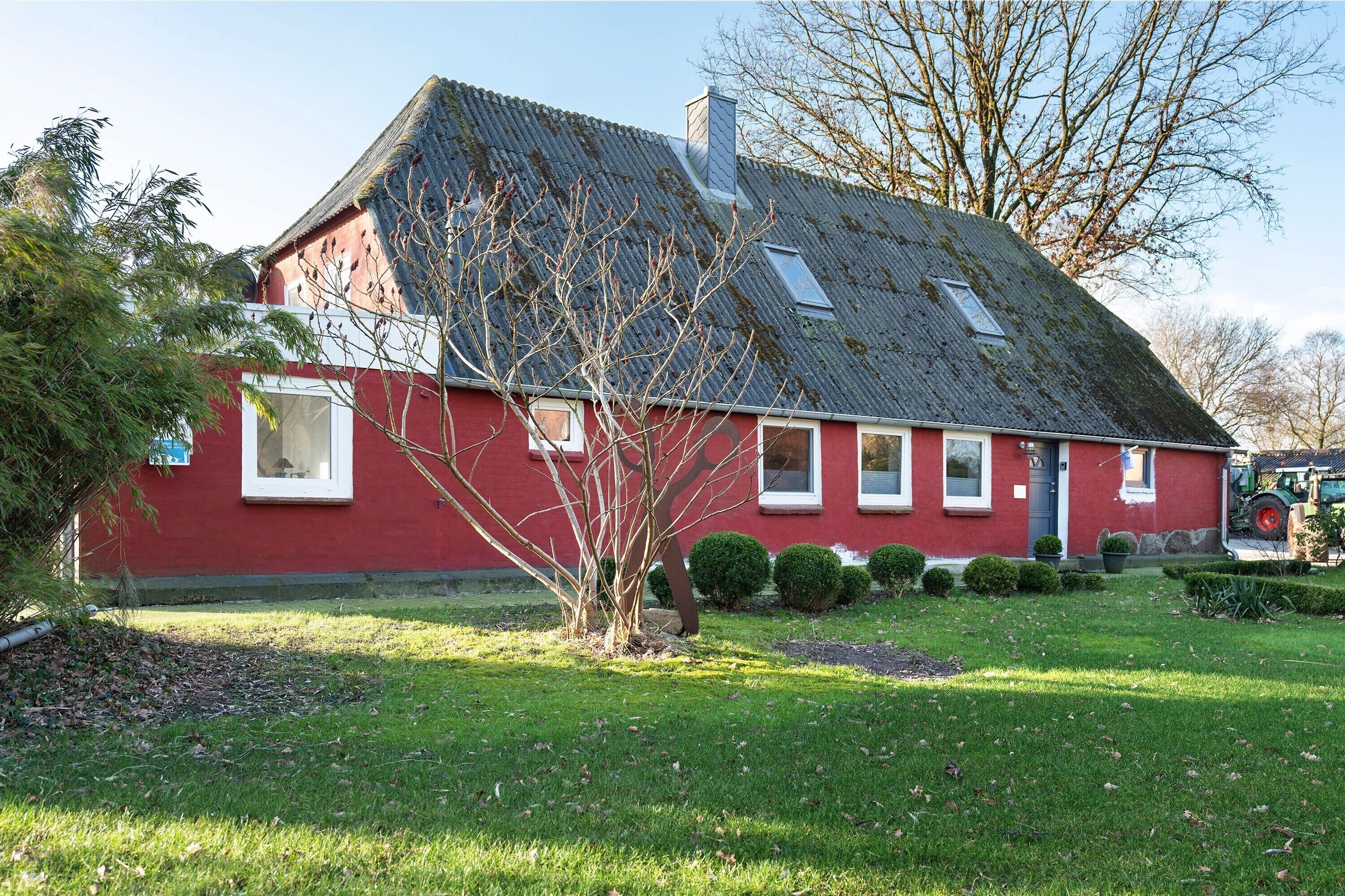 Een rood gebouw met een schuin dak, schoorsteen en een kleine tuin met een boom en struiken.
