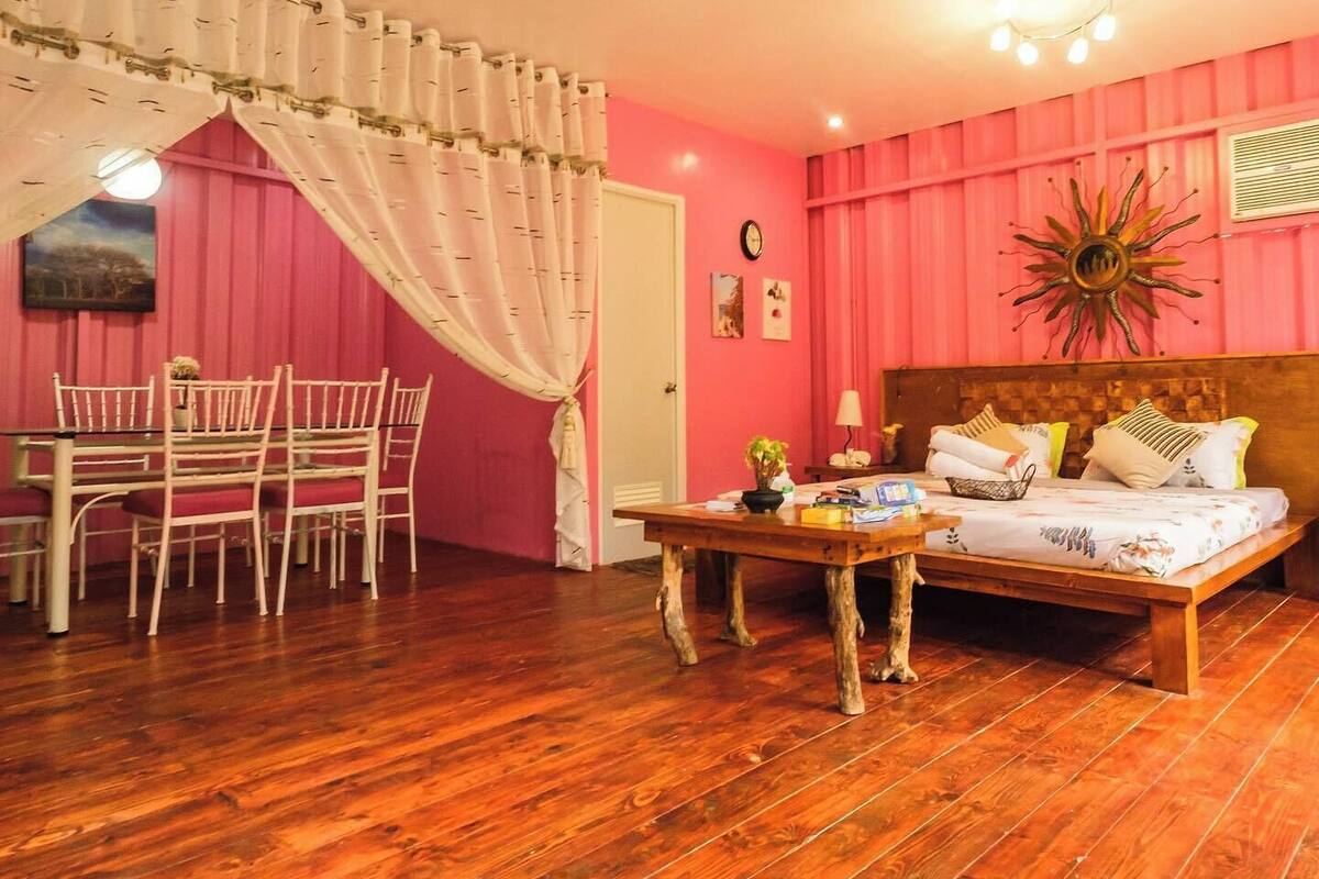 Una habitación con piso de madera, una cama con almohadas, una mesa con lámpara, una silla y una ventana con cortinas.