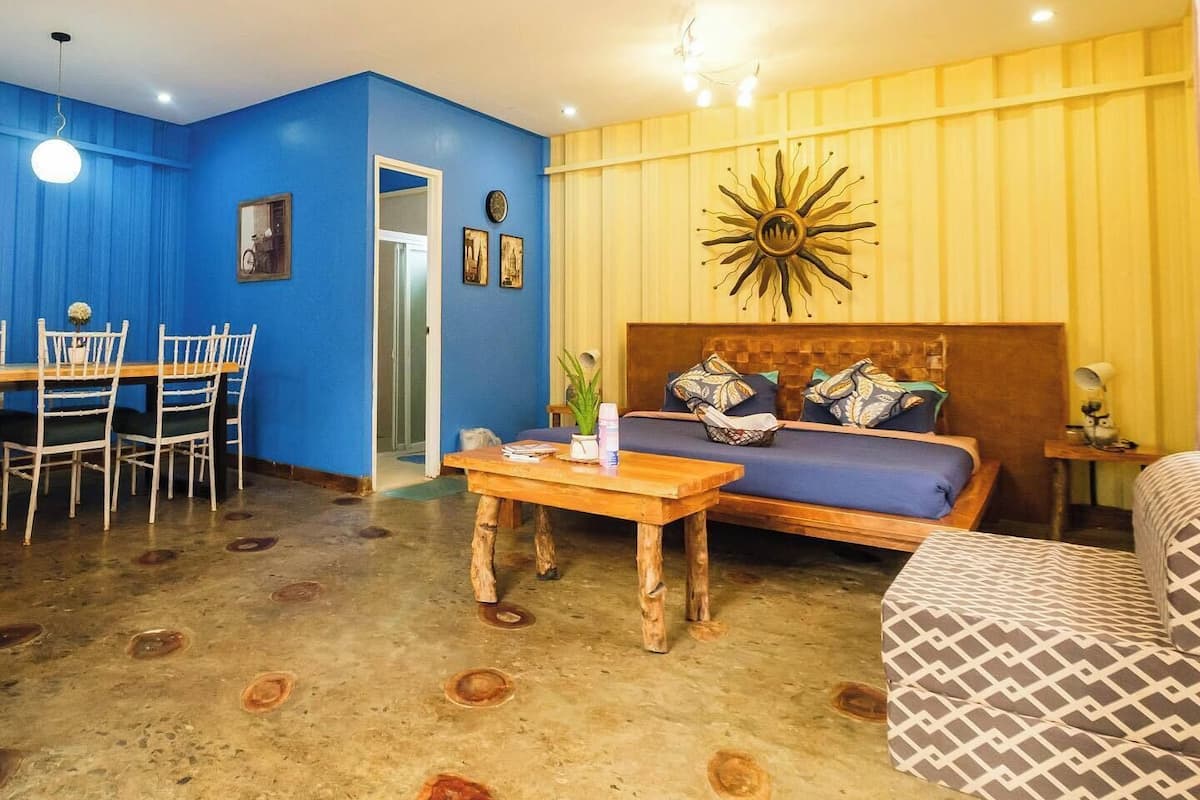Una habitación con pared azul, un sofá de madera con cojines estampados, una mesa de centro de madera y un área de comedor con una mesa y sillas.