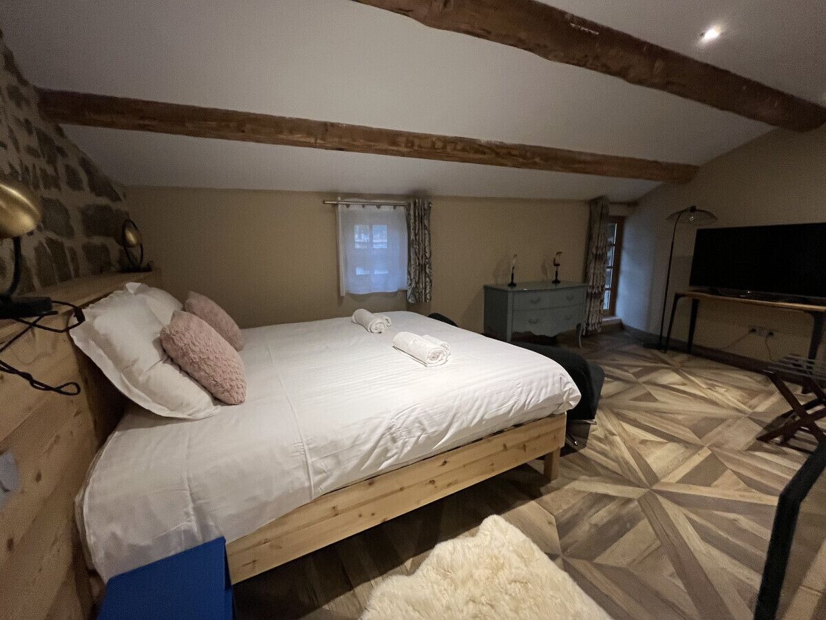 Quarto com cama de madeira, parede de pedra, janela com cortinas, escrivaninha e televisão.