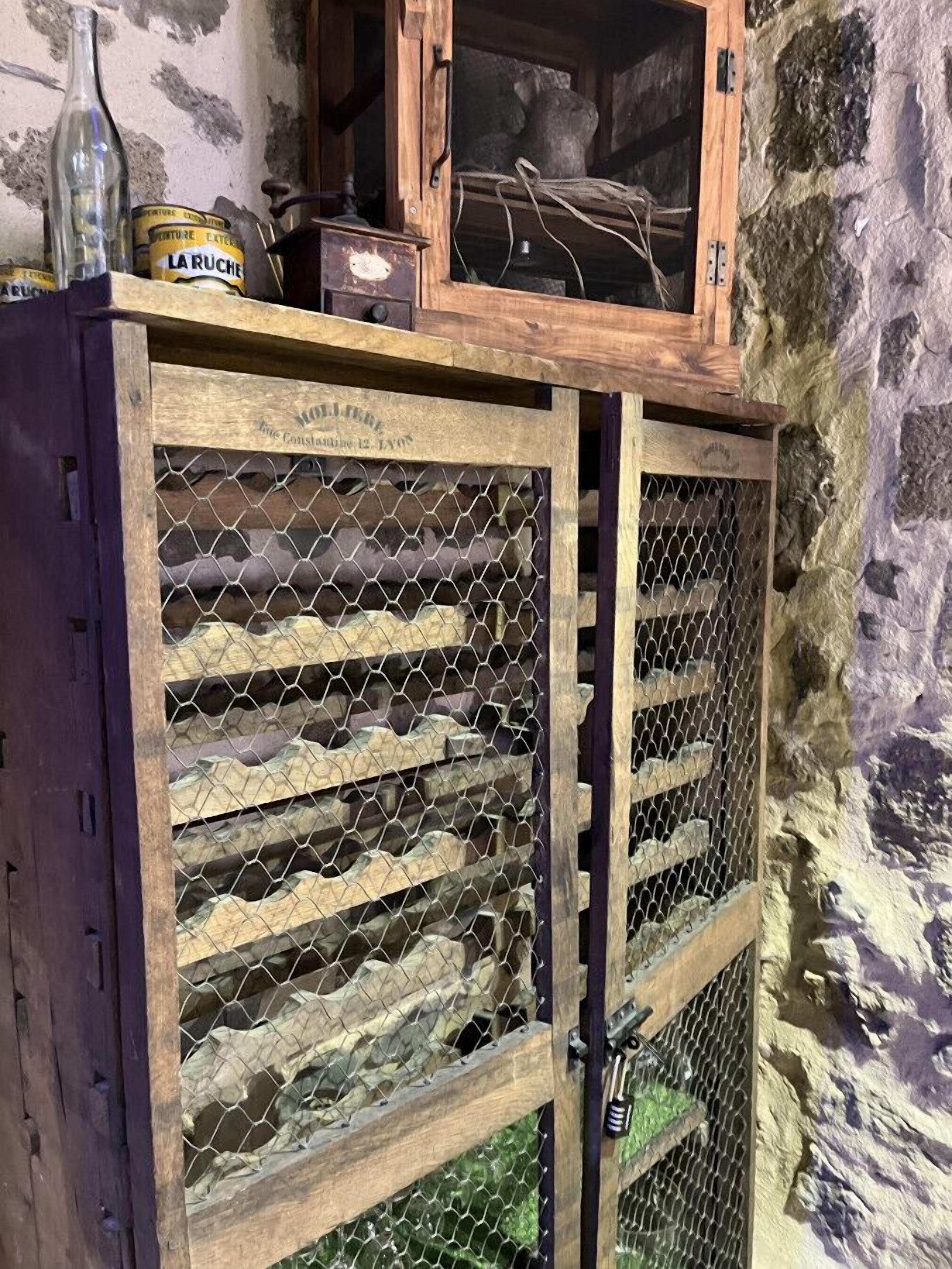 Een houten wijnkelder met een metalen hekwerkdeur, een fles Laruche wijn en een klein houten kistje.