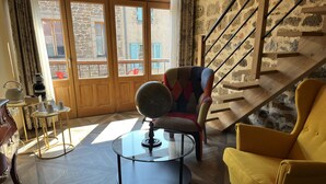 Una habitación con una mesa redonda de vidrio, un sillón amarillo, un sillón multicolores y una escalera con una pared de piedra.