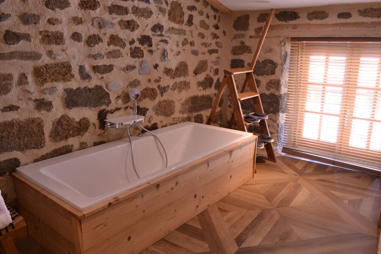Un baño con pared de piedra, una bañera de madera, una escalera de madera y una ventana con persianas.