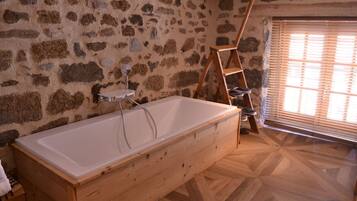 Un baño con pared de piedra, una bañera de madera, una escalera de madera y una ventana con persianas.