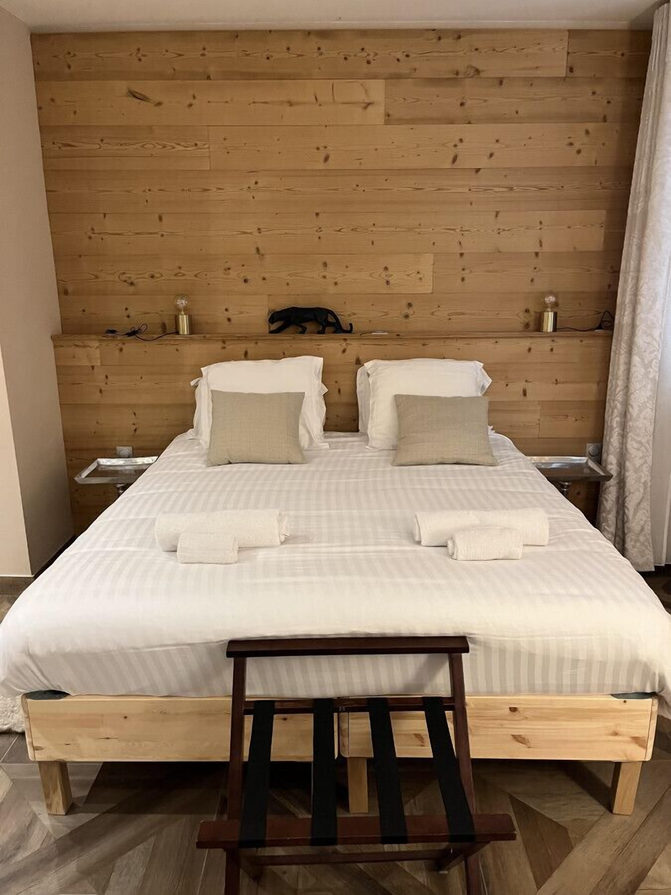 Un lit double avec une literie blanche et deux oreillers, une tête de lit en bois, une table de chevet avec une lampe, et un banc en bois placé devant le lit.