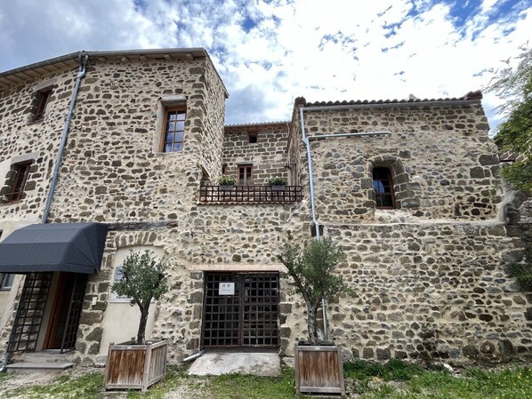 A stone building with a balcony, a door with a sign, and a small tree in a planter.