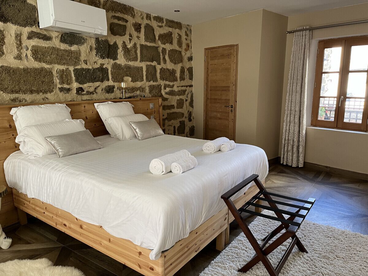 Un dormitorio con pared de piedra, estructura de cama de madera, ropa de cama blanca y una silla de madera.