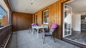 Un balcón de madera con una mesa, sillas y un ventanal corredizo, con vistas al exterior.