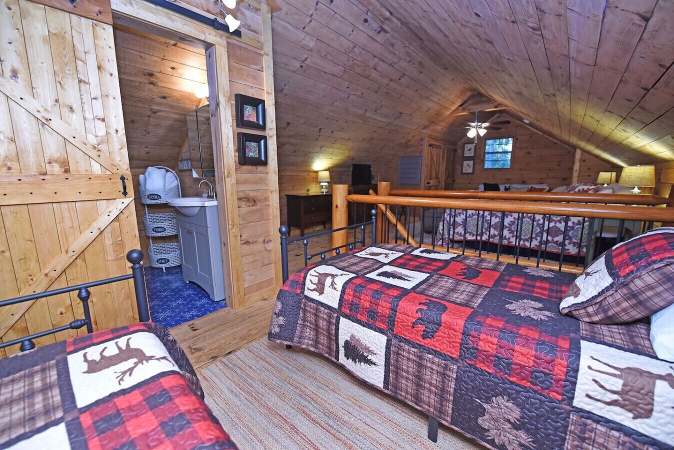 Une chambre mansardée chaleureuse avec un plafond en bois, un lit recouvert d’une courtepointe à motifs animaliers, un petit bureau avec une chaise et une fenêtre laissant entrer la lumière naturelle.