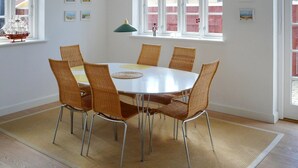 Een eetruimte met een ronde tafel en zes rieten stoelen, grote ramen met witte kaders en een hangende kroonluchter.