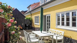 Een terras buiten met een witte tafel en stoelen, een groene parasol en een geel gebouw op de achtergrond.