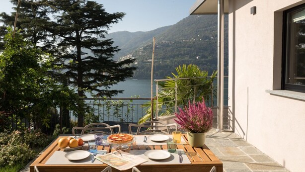 A table set outdoors with a view of a lake and mountains, surrounded by greenery. - VIlla Olivo - MyHomeinComo (Blevio)