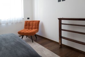 Une pièce comprenant un lit, un fauteuil orange, une table d’appoint en bois et une fenêtre ornée de rideaux légers.
