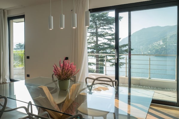 Un espace de restauration moderne avec une table en verre, des chaises blanches, un vase rempli de fleurs roses et une vue sur un lac et des montagnes, visible à travers de grandes fenêtres.