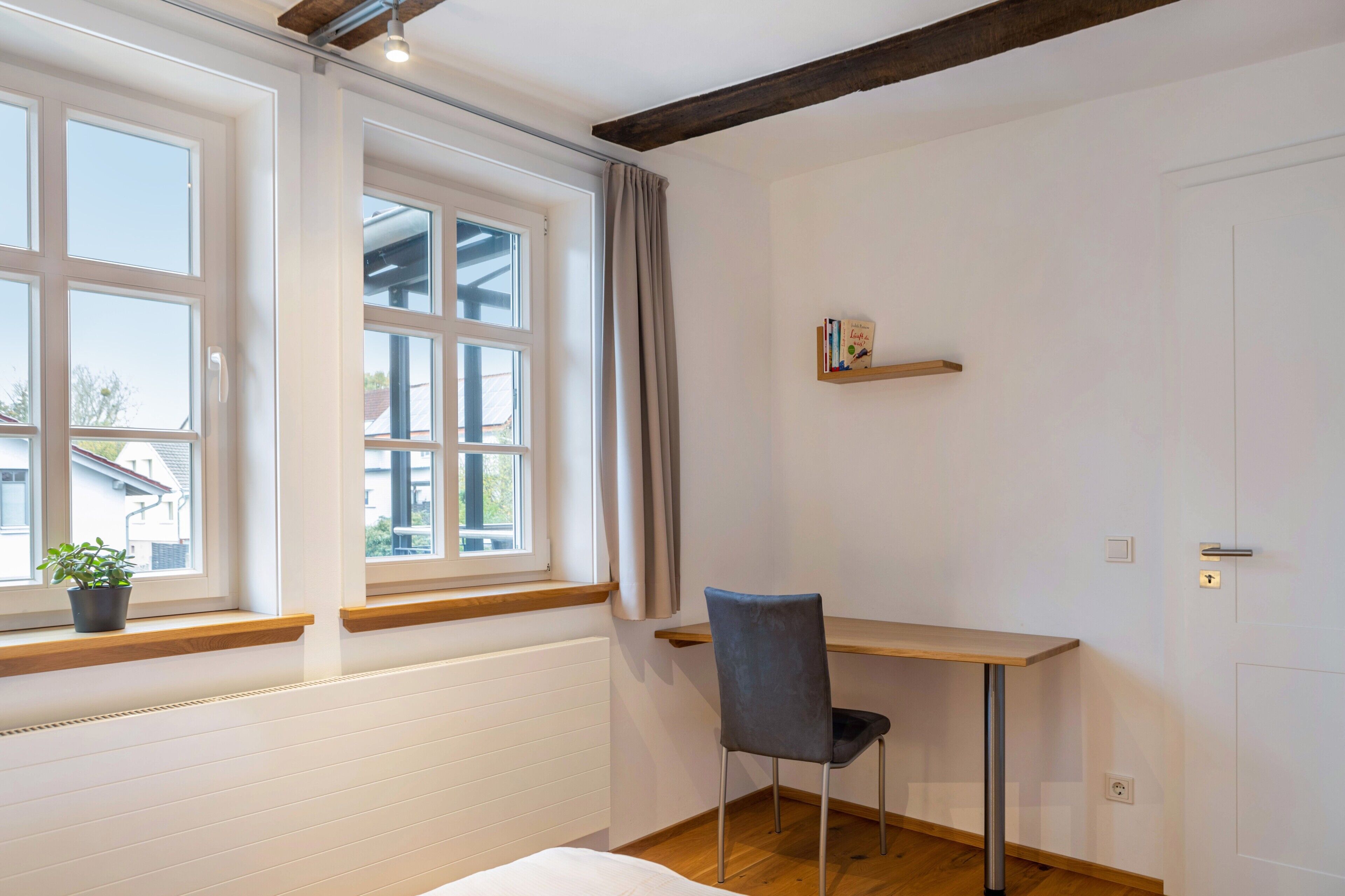 Een kamer met een bed, een bureau, een stoel, een raam met gordijnen, een radiator en een klein plankje met boeken.