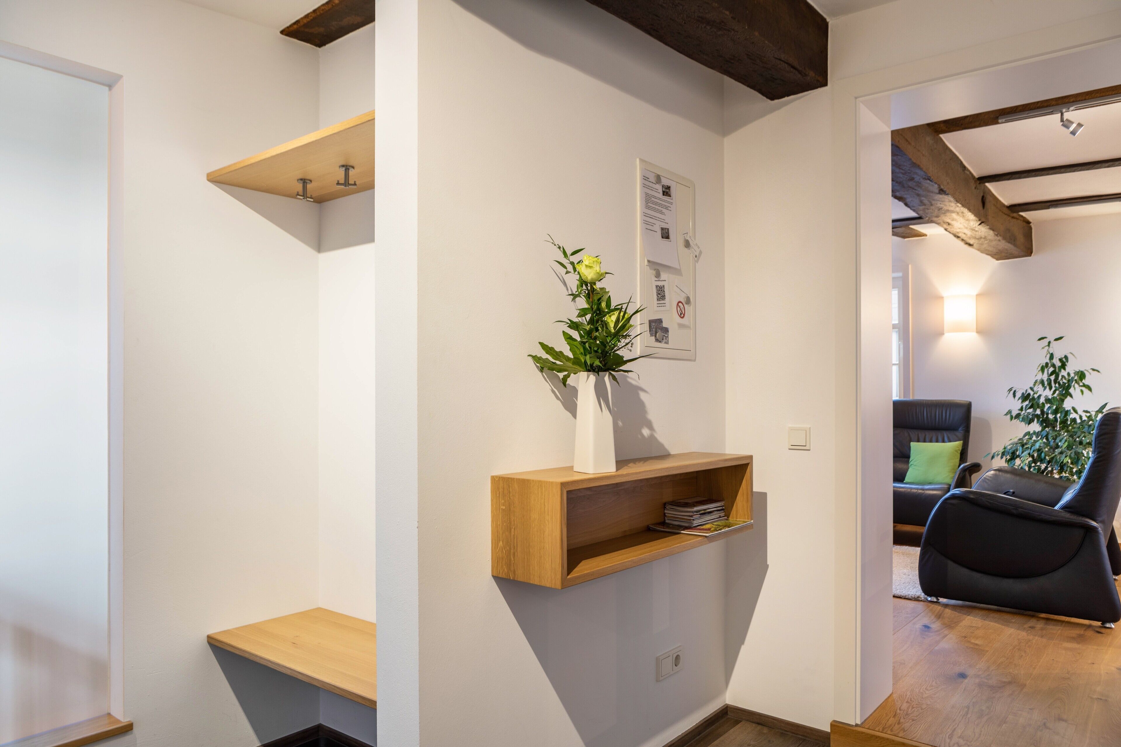 Un intérieur moderne avec un banc en bois, une étagère murale ornée d’un vase et un coin salon comprenant une chaise noire et une plante.