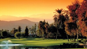 Un campo de golf con un estanque, árboles y montañas a lo lejos.