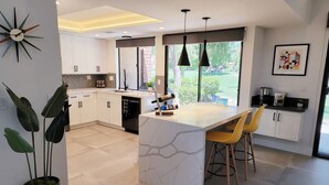 Una cocina moderna con armarios blancos, una isla de mármol, sillas amarillas, lámparas colgantes y un amplio ventanal con vistas al exterior.