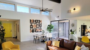 Amplia sala de estar con un sofá de cuero marrón, un sillón amarillo y una mesa de comedor con sillas. Cuenta con un gran ventanal que proporciona luz natural y un ventilador de techo.