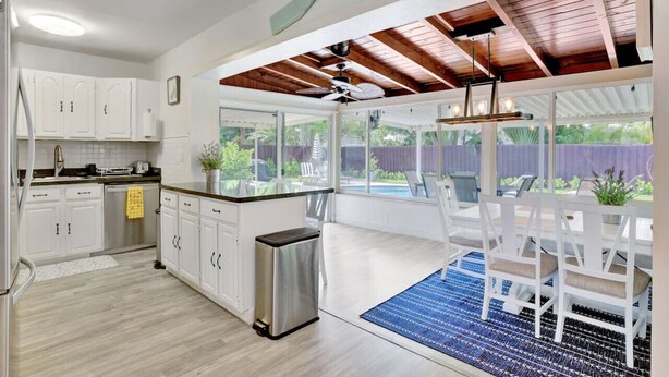 A kitchen with white cabinets, a stainless steel trash can, and a dining area with white chairs and a blue rug. - Eagle`s Way: Heated Pool, Firepit & Sunroom Near BEACH! (North Palm Beach)