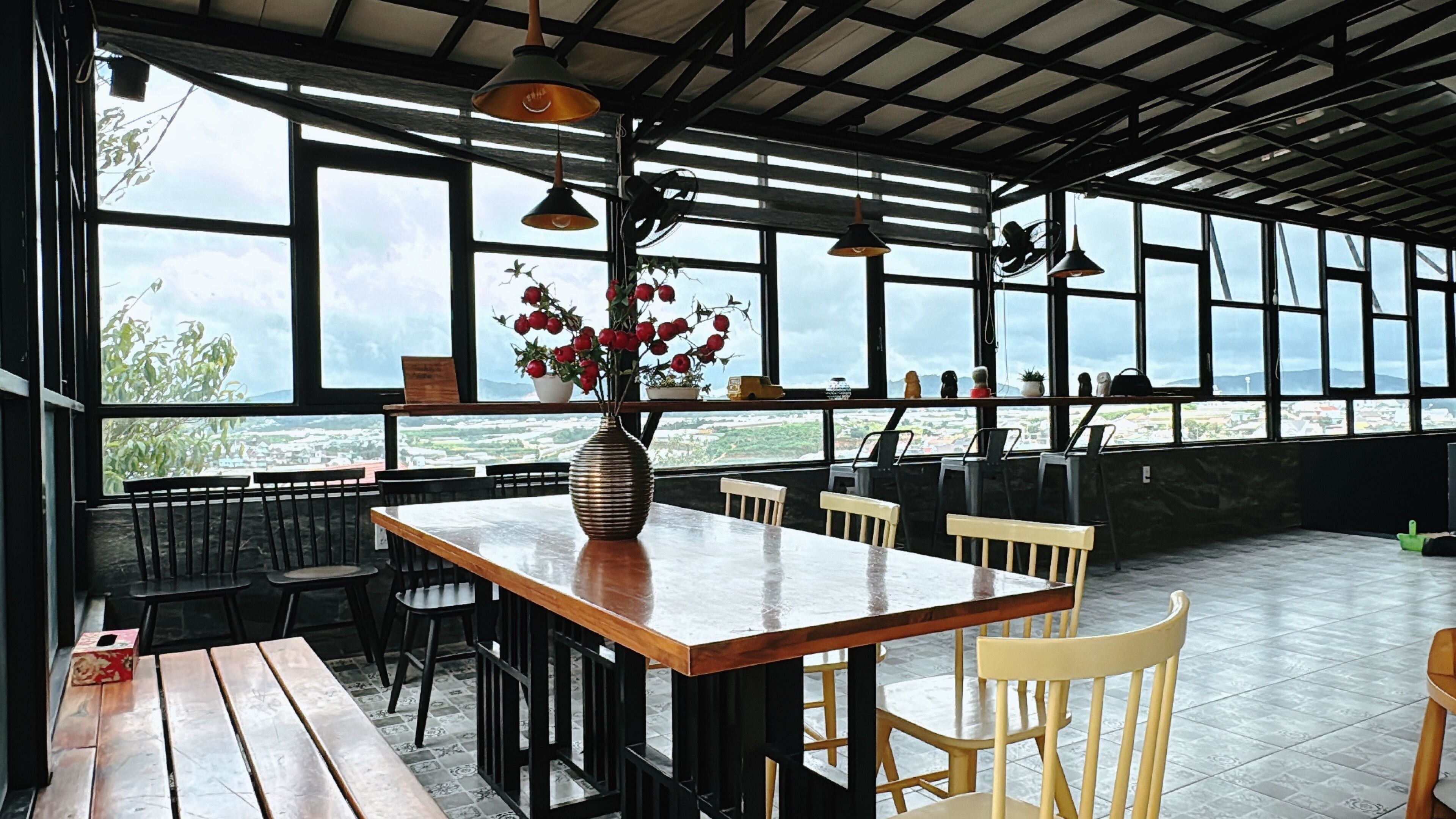 A restaurant with a large wooden table, black and white chairs, and a vase of red flowers. Large windows provide a view of the city and a body of water.