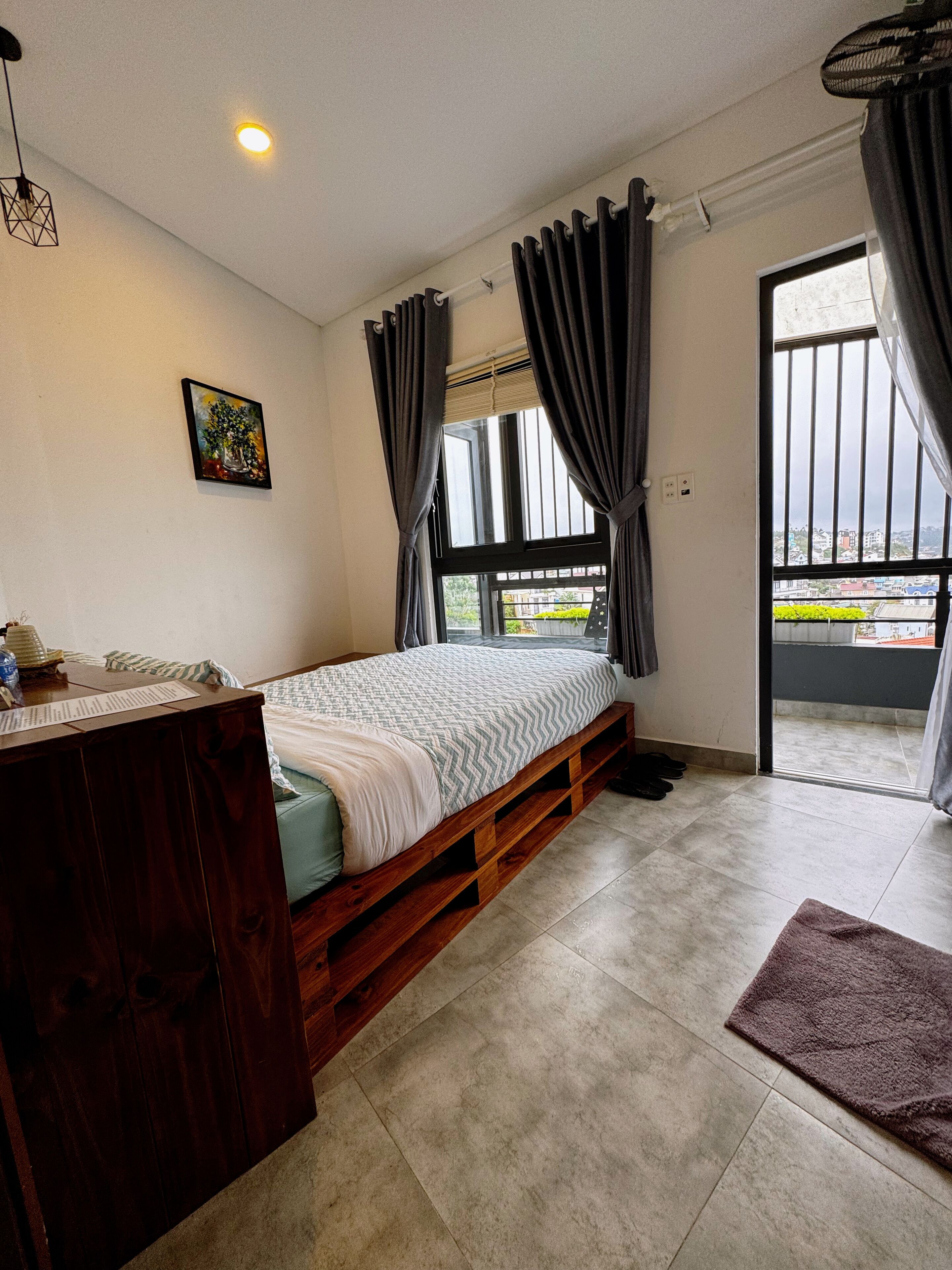 Un dormitorio con cama, escritorio de madera, ventana con cortinas, un cuadro en la pared y vistas al exterior.