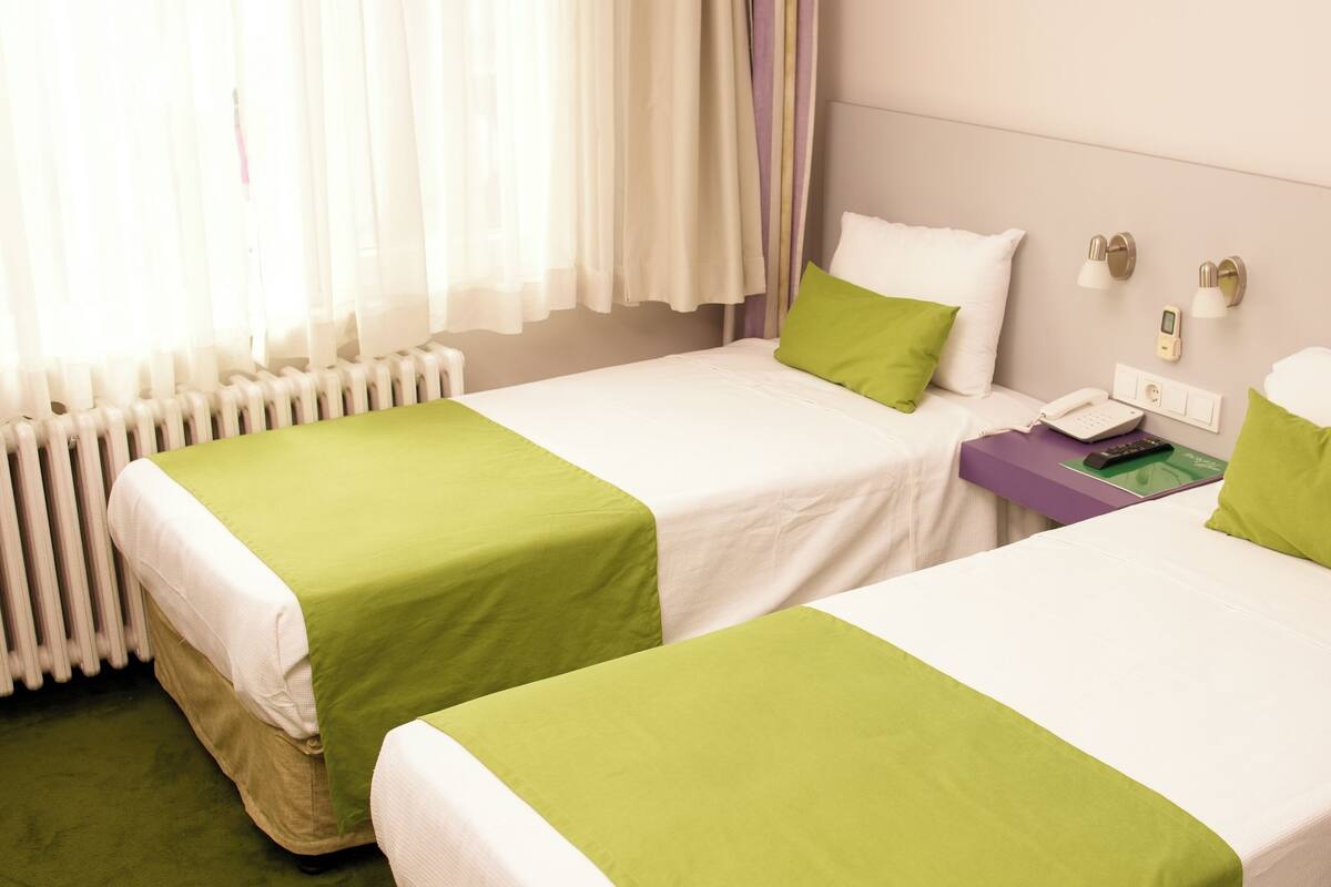 Hotellihuone, jossa on kaksi sänkyä, ja molemmissa on vihreät ja valkoiset vuodevaatteet. Huoneessa on yöpöytä, jossa on puhelin ja kaukosäädin, sekä ikkuna, jossa on läpikuultavat verhot.