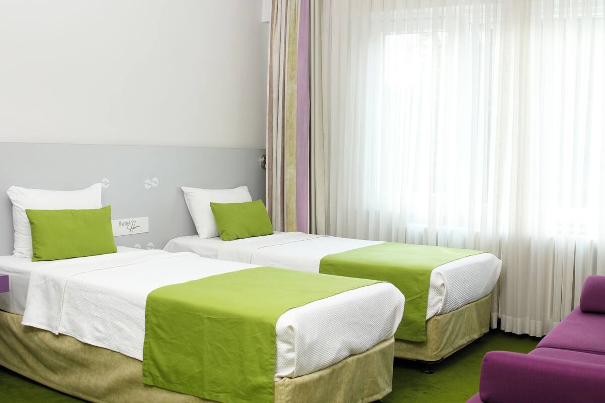 Hotellihuone, jossa on kaksi sänkyä, ja molemmissa on vihreät ja valkoiset vuodevaatteet. Huoneessa on myös violetti tuoli ja ikkuna, jossa on läpikuultavat verhot.