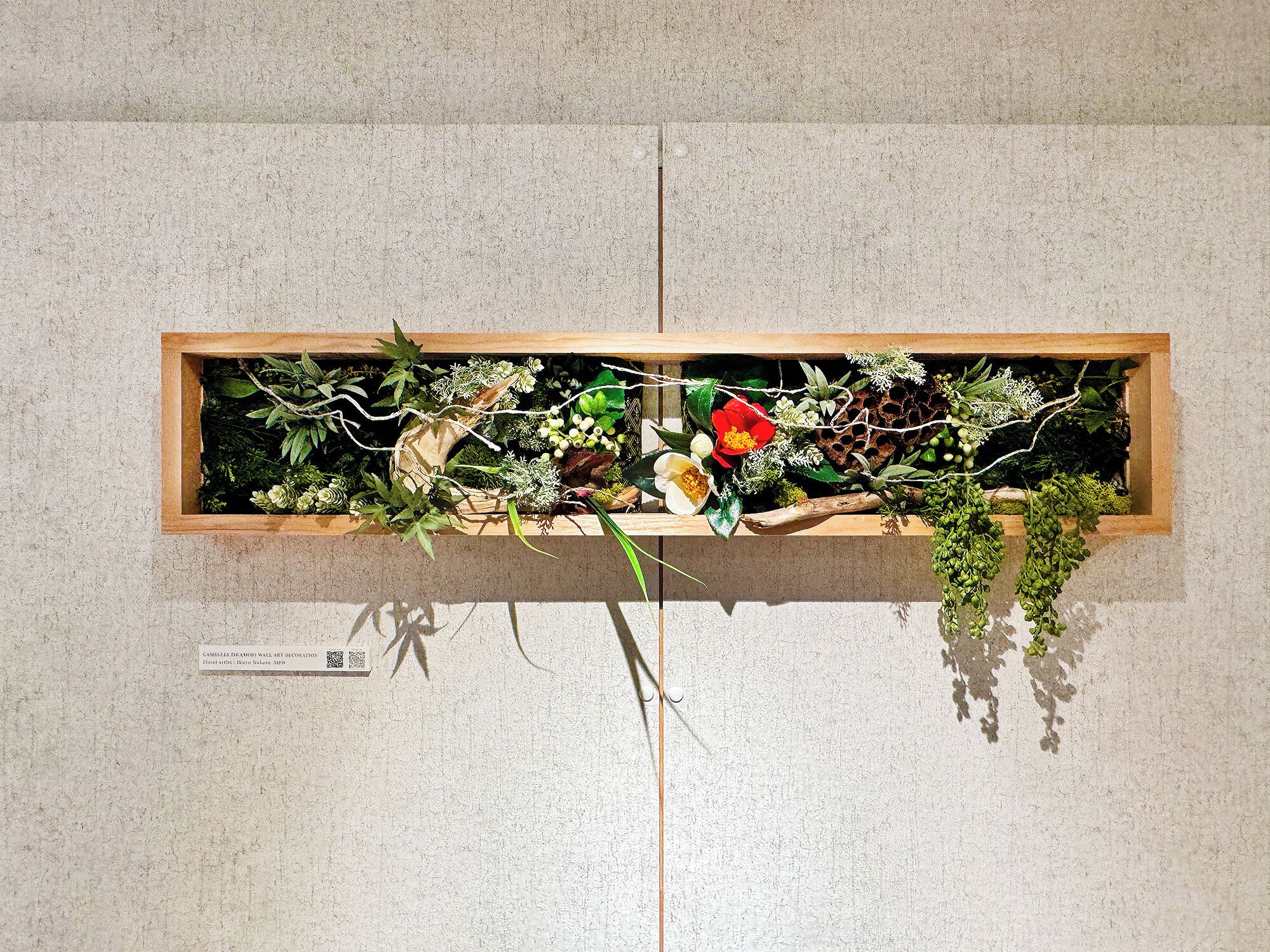 A wooden planter box with various plants and flowers against a textured wall.