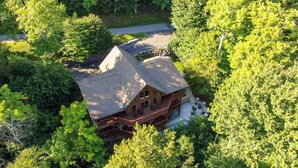 A large log cabin with a prominent gabled roof, surrounded by dense forest. - One Day More (Banner Elk)