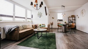A living room with a sofa, coffee table, and dining area with chairs. There is a window with blinds, a chandelier, and a wall-mounted shelf with decorative items. - Apartment 'Ferienwohnung Emma' with Mountain View and Wi-Fi (Güntersberge)