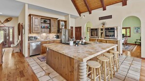 Une cuisine spacieuse avec des meubles en bois, un grand îlot central doté de tabourets, des appareils électroménagers en acier inoxydable et un plafond haut avec des poutres apparentes.