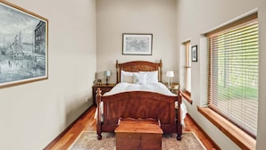 Une chambre avec un lit en bois, une table de chevet, une commode, une fenêtre avec des stores, un tableau encadré au mur et un tapis sur le sol.